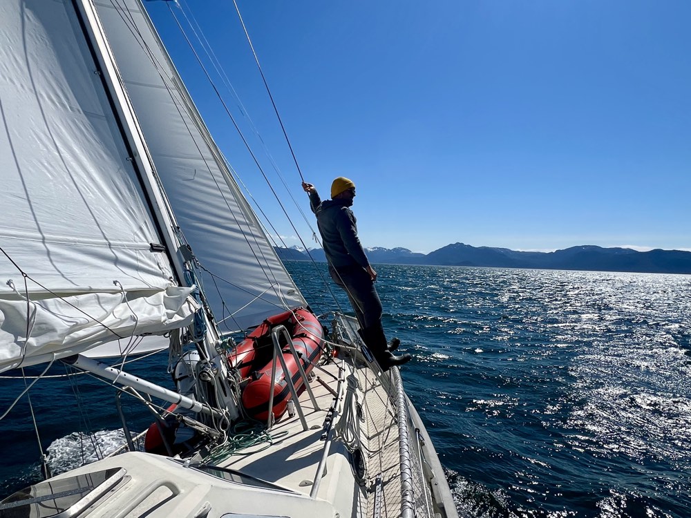 Sailor on the foredeck of Mazu II under sail, Alaskan mountains on the horizon